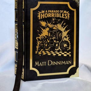 Puede incluir: Un libro de tapa dura negro titulado A Parade of Horribles de Matt Dinniman, con ilustraciones en l&aacute;mina dorada de un veh&iacute;culo y personajes en la portada, esquinas doradas y un marcap&aacute;ginas de cinta marr&oacute;n oscuro con un peque&ntilde;o dije de calavera.