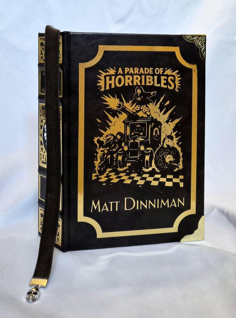 Puede incluir: Un libro de tapa dura negro titulado A Parade of Horribles de Matt Dinniman, con ilustraciones en l&aacute;mina dorada de un veh&iacute;culo y personajes en la portada, esquinas doradas y un marcap&aacute;ginas de cinta marr&oacute;n oscuro con un peque&ntilde;o dije de calavera.