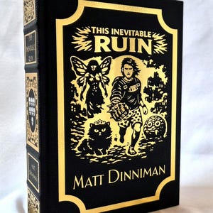Puede incluir: Un libro de tapa dura negro titulado This Inevitable Ruin de Matt Dinniman. La portada presenta ilustraciones en l&aacute;mina de oro de una figura alada, una persona con un guantelete de p&uacute;as, un gato con corona y una peque&ntilde;a criatura.