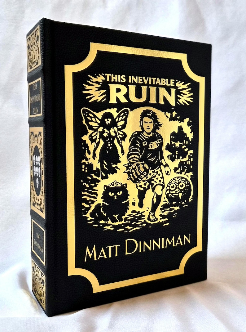 Puede incluir: Un libro de tapa dura negro titulado This Inevitable Ruin de Matt Dinniman. La portada presenta ilustraciones en l&aacute;mina de oro de una figura alada, una persona con un guantelete de p&uacute;as, un gato con corona y una peque&ntilde;a criatura.
