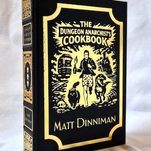 Puede incluir: Un libro de tapa dura negro titulado The Dungeon Anarchist's Cookbook de Matt Dinniman. La portada presenta un estampado de l&aacute;mina dorada con una ilustraci&oacute;n central de una persona corriendo con un gato y un dinosaurio, flanqueada por un tren y un autob&uacute;s.