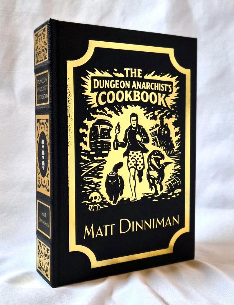 Puede incluir: Un libro de tapa dura negro titulado The Dungeon Anarchist's Cookbook de Matt Dinniman. La portada presenta un estampado de l&aacute;mina dorada con una ilustraci&oacute;n central de una persona corriendo con un gato y un dinosaurio, flanqueada por un tren y un autob&uacute;s.