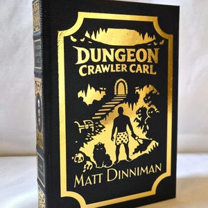 Puede incluir: Un libro de tapa dura negro titulado Dungeon Crawler Carl de Matt Dinniman. La portada presenta un estampado de l&aacute;mina dorada que representa a una figura en calzoncillos estampados en una mazmorra con un gato, un cofre y monstruos bajo un arco.
