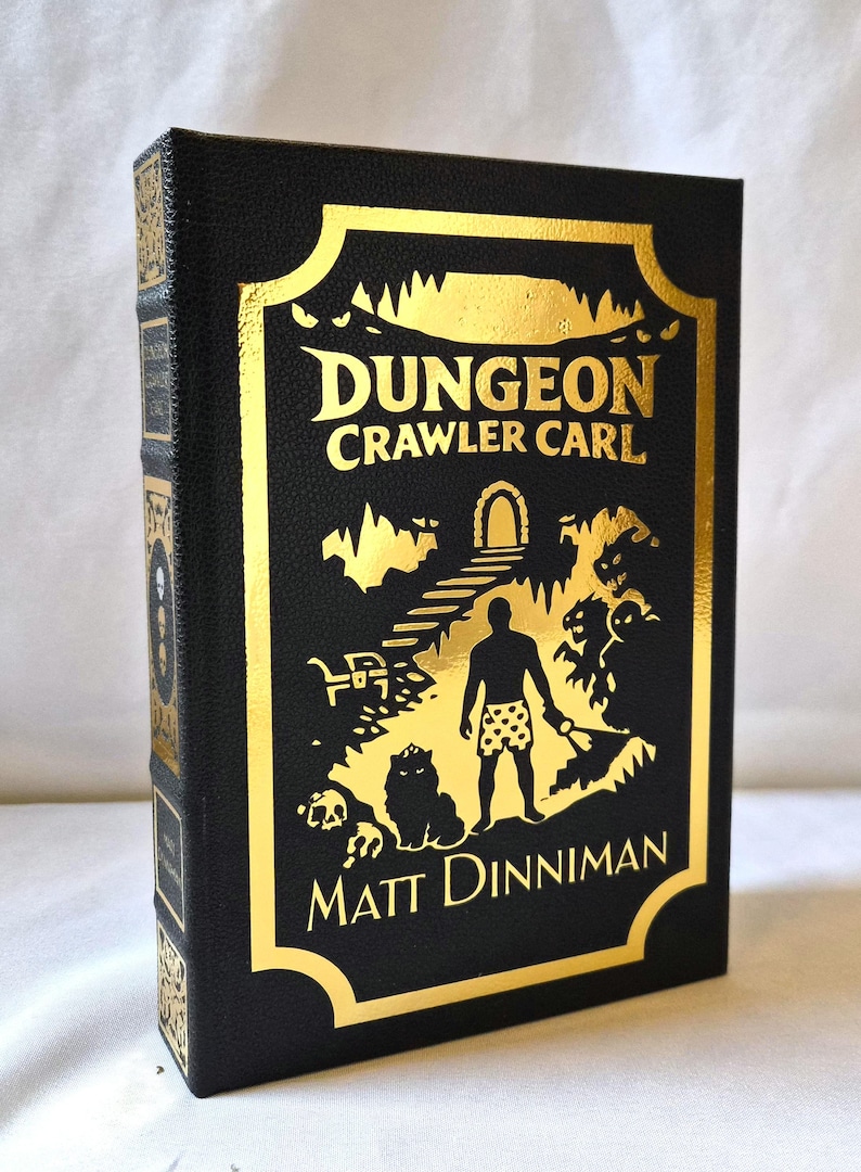 Puede incluir: Un libro de tapa dura negro titulado Dungeon Crawler Carl de Matt Dinniman. La portada presenta un estampado de l&aacute;mina dorada que representa a una figura en calzoncillos estampados en una mazmorra con un gato, un cofre y monstruos bajo un arco.