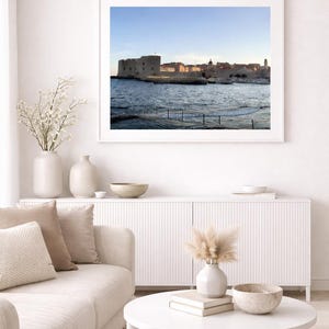 Könnte beinhalten: Ein gerahmtes Foto einer Küstenstadt mit Steingebäuden und einer Festung am Wasser. Das Bild wird über einem weißen Schrank und einem cremefarbenen Sofa mit dekorativen Kissen angezeigt. Ein weißer Couchtisch enthält eine Vase, Bücher und eine dekorative Schale.