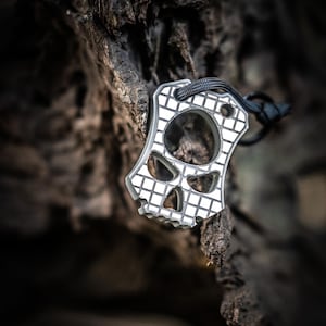 May include: A silver-colored, skull-shaped keychain with a grid pattern and a black lanyard. The keychain has cut-out eye and nose holes. The background is a blurred, dark brown, textured surface.