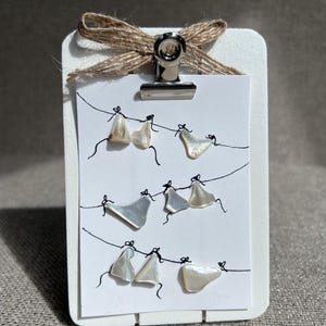 May include: A white rectangular display board with a silver metal clip holding a small white card. The card features hand-drawn black lines resembling clotheslines with small, iridescent, off-white shell fragments attached to the lines.