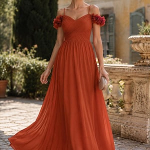 Könnte beinhalten: Ein fließendes, rostfarbenes Abendkleid mit Spaghettiträgern und floralen Schulterakzenten. Das Kleid hat ein plissiertes Oberteil und einen bodenlangen Rock. Eine kleine, goldene Clutch wird gehalten.