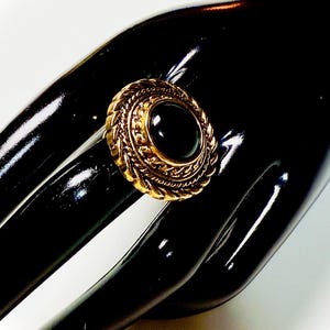 Puede incluir: Un anillo dorado con una gran piedra negra ovalada. El anillo tiene un diseño detallado y texturizado alrededor de la piedra, con un borde trenzado. El anillo está sobre un fondo negro.