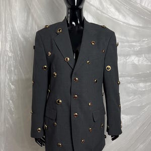 May include: A dark gray blazer with numerous gold-colored buttons and decorative eyelets. The blazer has a classic double-breasted design with two front pockets. The garment is displayed on a black mannequin.