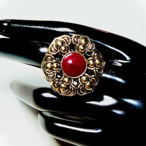 Puede incluir: Un anillo dorado con una piedra central redonda y de color rojo intenso. El anillo presenta un diseño ornamental de inspiración floral alrededor de la piedra. El anillo se muestra sobre una superficie negra, creando un contraste llamativo.