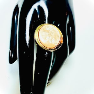 Puede incluir: Un anillo dorado con un gran centro redondo de color crema. El centro del anillo tiene un aspecto veteado. El anillo se muestra sobre un fondo negro, creando un contraste que resalta su diseño.