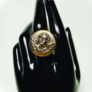Puede incluir: Anillo dorado con un diseño detallado de querubín. El anillo es redondo y está engarzado en una mano negra, creando un contraste llamativo. El querubín tiene alas y rasgos faciales detallados.