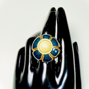 Puede incluir: Un anillo dorado con diseño de flor. El anillo presenta una perla central de color crema rodeada de pétalos de esmalte azul. El anillo está sobre una superficie negra.