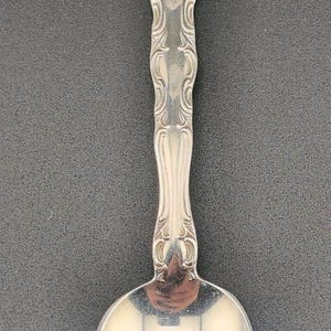 Può includere: Un cucchiaio d'argento con un manico ornato e una coppa arrotondata. Il manico presenta un design simmetrico dettagliato con motivi a volute. Il cucchiaio è probabilmente in argento sterling ed è mostrato su uno sfondo scuro.