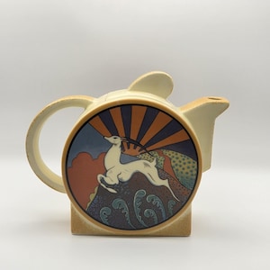 May include: A cream-coloured ceramic teapot featuring a circular central panel with a graphic illustration of a leaping gazelle. The background depicts a stylised sunburst in orange and dark blue, with textured landscape elements in muted earth tones.