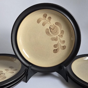 May include: A collection of beige ceramic plates with dark brown rims. Each plate has a brown floral pattern. The plates are displayed on a black stand, highlighting their decorative appeal. Ideal for adding a touch of elegance to a dining setting.
