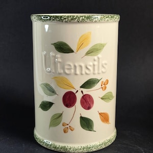 May include: Cream ceramic utensil holder with embossed "Utensils" text. Decorated with painted green, yellow, and orange leaves, and red cherries. The rim and base have a green speckled design. A kitchen decor item.