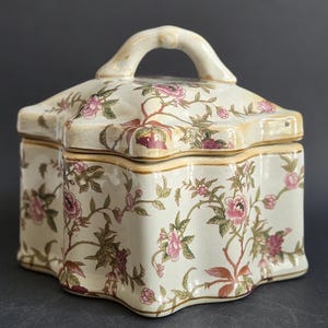 May include: A cream-coloured ceramic lidded box with a scalloped edge and a central handle on the lid. The surface is decorated with a repeating pattern of pink flowers, green leaves, and brown stems, featuring a glossy finish and gold trim.