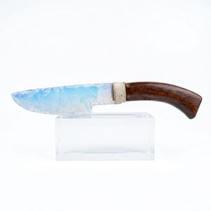 Puede incluir: Un cuchillo artesanal con una hoja opalina azul claro y un mango de madera marrón oscuro. La hoja está asegurada al mango con un cordón blanco roto enrollado. El cuchillo se exhibe sobre un soporte de acrílico transparente.