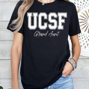 May include: Black t-shirt with the white text "UCSF Grand Aunt" in a collegiate font. The shirt is a crew neck style and is worn by a person wearing blue jeans.