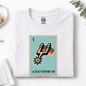 May include: White t-shirt with a graphic featuring a teal card with the number 1 and the text "LA CRAZY HISPANIC FAN". The graphic includes a lightning bolt, a starburst, and a flag with red, yellow, and green stripes.