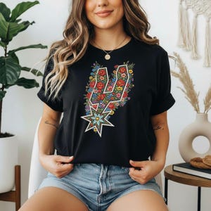 Może przedstawiać: Czarny t-shirt z kwiatowym wzorem w kształcie logo drużyny. Wz&oacute;r zawiera czerwone, niebieskie, ż&oacute;łte i zielone kwiaty. Koszulka ma okrągły dekolt i kr&oacute;tkie rękawy.