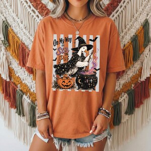 May include: An orange short-sleeve t-shirt featuring a graphic of a goose wearing a witch hat and cape, stirring a black cauldron. The text reads On My Way To Stir The Pot. The design includes a jack-o'-lantern, bats, and stars on a white background.