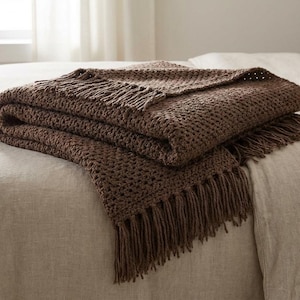 May include: A folded brown crochet blanket with a fringed edge. The blanket, made from thick yarn, is draped over a bed. The neutral colour and simple design make it a versatile home decor item, suitable for various interior styles.