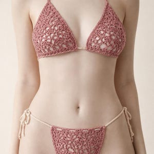 Könnte beinhalten: Gehäkeltes Bikini-Set in Staubrosa. Das Triangel-Oberteil und der String-Bikini-Slip haben ein offenes, spitzenartiges Design. Cremefarbene Bänder zum Binden. Das Set ist zart und handgefertigt, perfekt für den Strand.