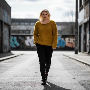 May include: A woman wearing a mustard yellow sweater and black pants walks down a city street. The sweater has a round neckline and long sleeves. The pants are fitted. The background features graffiti art on the walls.