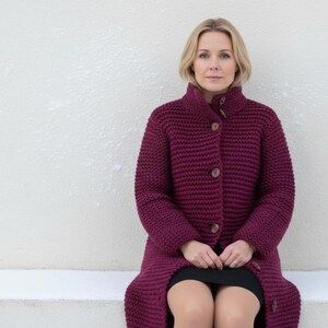 May include: A woman wearing a long, burgundy knit coat with large wooden buttons. The coat has a high collar and is knee-length. The woman is sitting in front of a white textured wall.