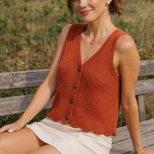 May include: A rust-colored, sleeveless, crocheted vest with a V-neck and scalloped hemline. The vest has four brown buttons down the front. The woman is wearing a white skirt and sitting on a wooden bench.