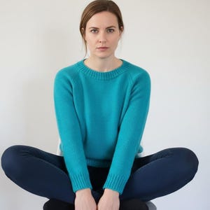 Könnte beinhalten: Eine Person trägt einen türkisfarbenen Rundhals-Pullover und dunkelblaue Jeans. Die Person sitzt mit überkreuzten Beinen vor einem weißen Hintergrund. Der Pullover hat gerippte Bündchen und einen Saum.