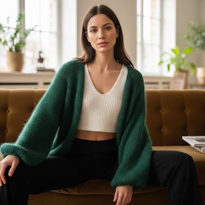 May include: A woman wearing a dark green, oversized cardigan with a white ribbed crop top and black pants. The cardigan has long sleeves. She is sitting on a brown sofa in a well-lit room.