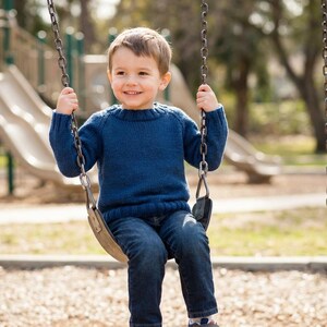 May include: A young child wearing a blue sweater and jeans sits on a swing set at a park. The child is smiling and holding onto the chains of the swing. A playground with slides is visible in the background.