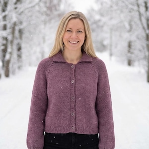 Könnte beinhalten: Eine Frau trägt einen mauvefarbenen Cardigan mit Knöpfen und Kragen. Der Cardigan ist gestrickt. Sie lächelt in einer verschneiten Umgebung im Freien mit Bäumen im Hintergrund. Der Cardigan hat einen Knopfverschluss.