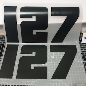Puede incluir: Un conjunto de dos calcomanías de vinilo negro de gran tamaño que muestran el número 127 en una tipografía sans-serif en negrita. Los números están dispuestos en dos filas sobre una hoja de transferencia transparente para su aplicación.