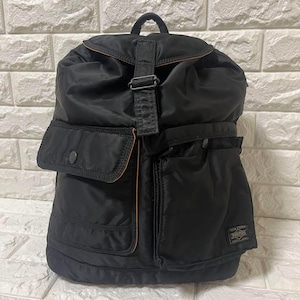 May include: A black nylon backpack featuring a top flap with a buckle closure, two front pockets with snap buttons, and a small rectangular label on the lower right corner with the text YOSHIDA & KANAME PORTER TOKYO JAPAN.