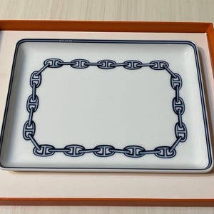 May include: A white rectangular tray with a blue chain link design around the border. The tray is inside an orange box. The word "HERMES" is printed in blue at the top of the tray.