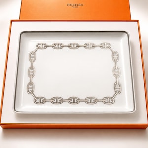 May include: A rectangular white porcelain tray featuring a silver-toned chain link pattern around the perimeter. The tray is presented inside an orange box with the text HERMES PARIS printed in gold on the interior lid of the box.