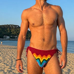 Puede incluir: Una braga de bikini de ganchillo color granate con un vibrante patrón de chevrón en amarillo, naranja, verde, azul y morado. La prenda incluye finos lazos laterales para un ajuste regulable y está confeccionada con tejido de punto.