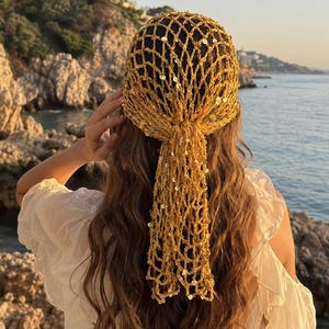 May include: A gold-colored crochet headscarf featuring an open net pattern embellished with small, reflective sequins. The headscarf is tied at the back of the head, with the ends hanging down in a loose, textured mesh design.