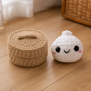 May include: A round, tan crocheted container with a matching lid sits beside a small, white, spherical crocheted plush toy. The plush features black plastic eyes, a stitched black smile, and small pink embroidered cheeks on its surface.