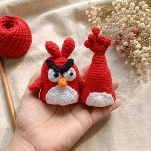 May include: Crocheted red Angry Birds character with black eyebrows, white belly, and orange beak. A matching red crocheted hat with a white base is next to the bird. A ball of red yarn is in the background.