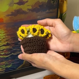 Puede incluir: Una maceta de girasoles hecha a mano con ganchillo. La maceta es marrón, con un interior blanco y rematada con girasoles amarillos. La artesanía se sostiene con dos manos. El fondo es una pintura de una puesta de sol sobre el océano.