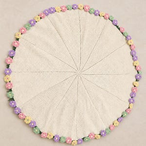 Peut inclure: Un napperon circulaire en crochet blanc cassé présentant un motif de points radiaux partant du centre. Le bord extérieur est fini avec une bordure décorative de petites fleurs en crochet multicolores dans des tons de rose, jaune, violet et vert.
