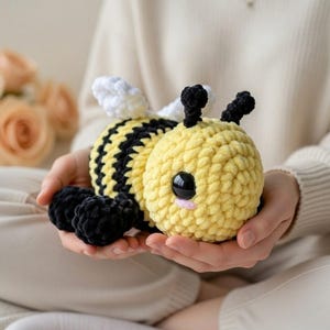 Peut inclure: Une peluche abeille faite à la main au crochet. L'abeille a un corps jaune avec des rayures noires, des antennes noires et de grands yeux noirs. Les ailes sont blanches. La peluche est tenue dans les mains de quelqu'un.