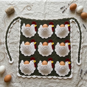 May include: A dark green crocheted egg holder featuring nine white chicken-shaped pockets with red combs and yellow beaks. Each pocket holds a brown egg. The rectangular piece has a white scalloped border and two long crocheted ties on the sides.