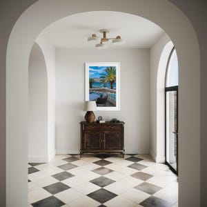 May include: An interior hallway with a black and white checkered floor, leading to a framed picture of a tropical scene. A dark wood cabinet with a lamp and a clock sits below the picture. The hallway is framed by white archways.